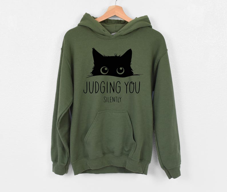 May include: A green hooded sweatshirt with a black cat peeking out from behind the text "JUDGING YOU SILENTLY".