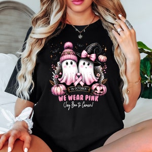 Op de afbeelding: Zwart T-shirt met een ontwerp van twee cartoongeesten met roze hoeden en een roze lint, met pompoenen en vleermuizen. De tekst luidt "In October We Wear Pink Say Boo to Cancer!"