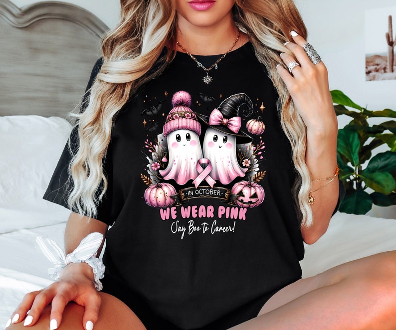 Op de afbeelding: Zwart T-shirt met een ontwerp van twee cartoongeesten met roze hoeden en een roze lint, met pompoenen en vleermuizen. De tekst luidt "In October We Wear Pink Say Boo to Cancer!"