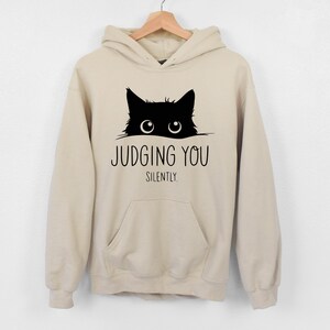 May include: A beige hoodie with a black cat peeking out from the top and the text "JUDGING YOU SILENTLY."