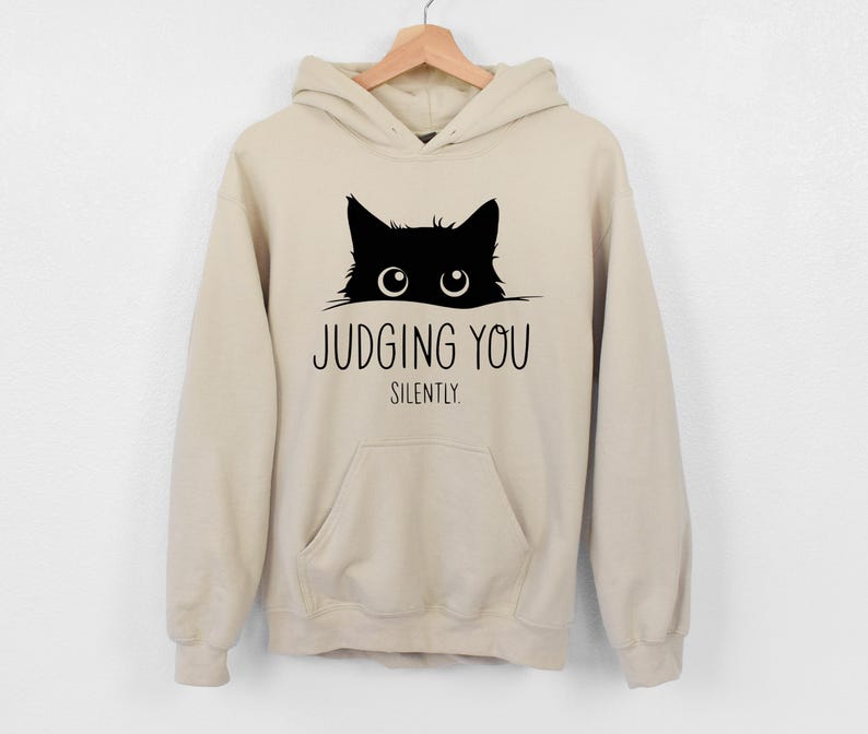 May include: A beige hoodie with a black cat peeking out from the top and the text "JUDGING YOU SILENTLY."