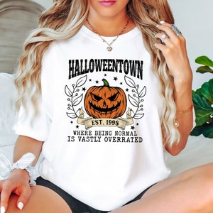 Puede incluir: Camiseta blanca con un dise&ntilde;o de Halloween. El dise&ntilde;o presenta una calabaza tallada, las palabras "Halloweentown" y "Est. 1998", y la frase "Where being normal is vastly overrated". El dise&ntilde;o est&aacute; rodeado de hojas y estrellas.