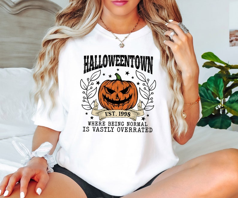 Puede incluir: Camiseta blanca con un dise&ntilde;o de Halloween. El dise&ntilde;o presenta una calabaza tallada, las palabras "Halloweentown" y "Est. 1998", y la frase "Where being normal is vastly overrated". El dise&ntilde;o est&aacute; rodeado de hojas y estrellas.