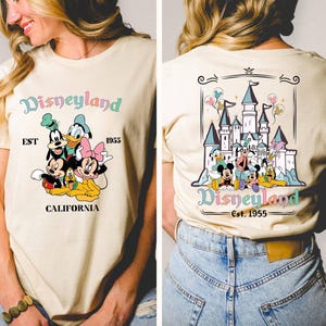 May include: A beige t-shirt with a graphic print of Mickey Mouse and friends in front of the Disneyland castle. The text "Disneyland Est. 1955 California" is printed on the shirt.
