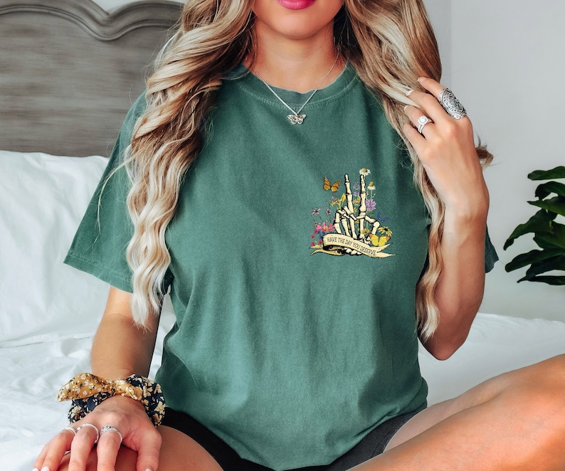 K&ouml;nnte beinhalten: Gr&uuml;nes T-Shirt mit einer Skeletthand, die ein Peace-Zeichen macht, umgeben von Blumen und Schmetterlingen. Der Text auf dem Shirt lautet "Have the day you deserve".