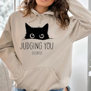 May include: A beige hoodie with a black cat peeking over the top and the text "JUDGING YOU SILENTLY."