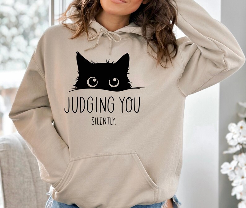 May include: A beige hoodie with a black cat peeking over the top and the text "JUDGING YOU SILENTLY."