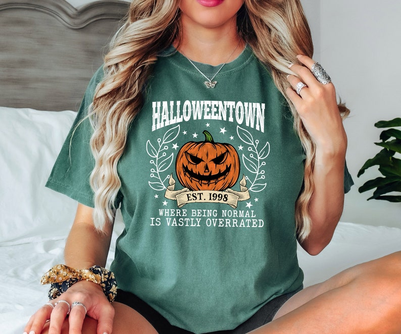 Puede incluir: Camiseta verde con el texto "Halloweentown" y un dise&ntilde;o de calabaza. El dise&ntilde;o incluye el texto "Est. 1998" y la frase "Where being normal is vastly overrated."