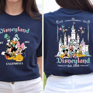 May include: A navy blue t-shirt with a graphic of Mickey Mouse and friends in front of Disneyland. The text "Disneyland Est. 1955 California" is printed on the shirt.