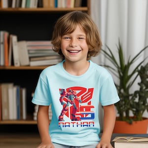 May include: A light blue t-shirt featuring a red and orange robot graphic with the name "NATHAN" in blue and white block letters. The shirt is worn by a smiling child.