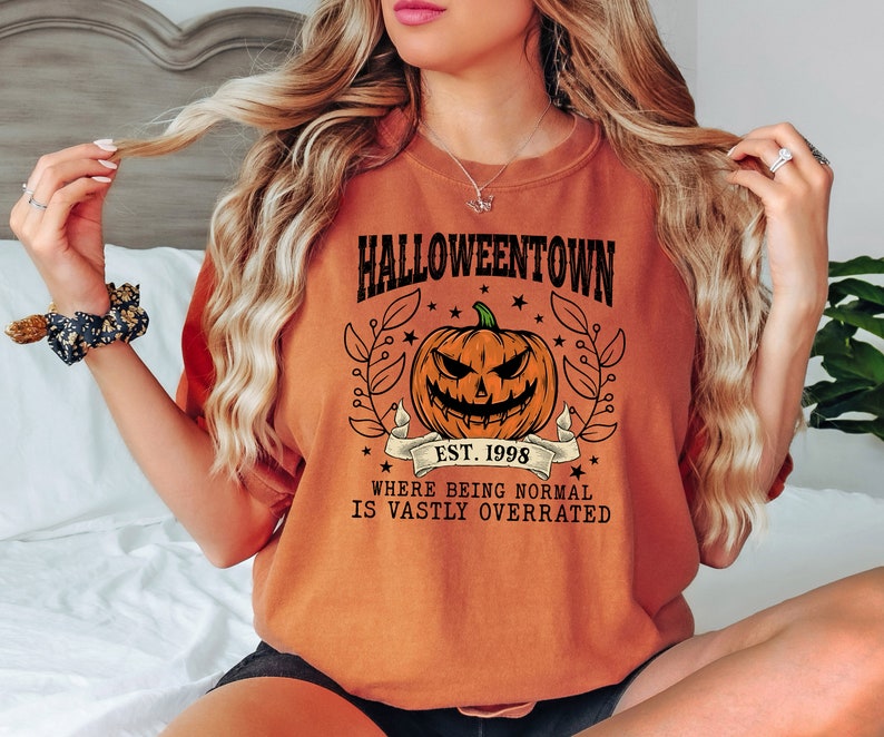Puede incluir: Camiseta naranja con un dise&ntilde;o de Halloween. El dise&ntilde;o presenta una calabaza tallada, el texto "Halloweentown Est. 1998" y la frase "Where being normal is vastly overrated."