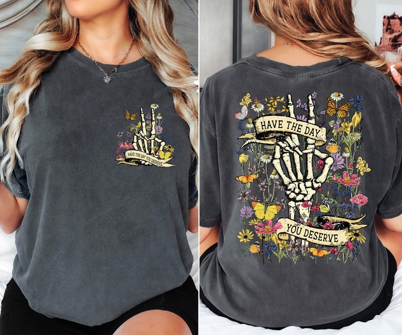 K&ouml;nnte beinhalten: Ein dunkelgraues T-Shirt mit einem Skelett-Hand-Motiv und dem Text "Have the day you deserve" auf der R&uuml;ckseite. Die Vorderseite des Shirts hat eine kleinere Version des Motivs mit dem Text "Have the day you deserve".