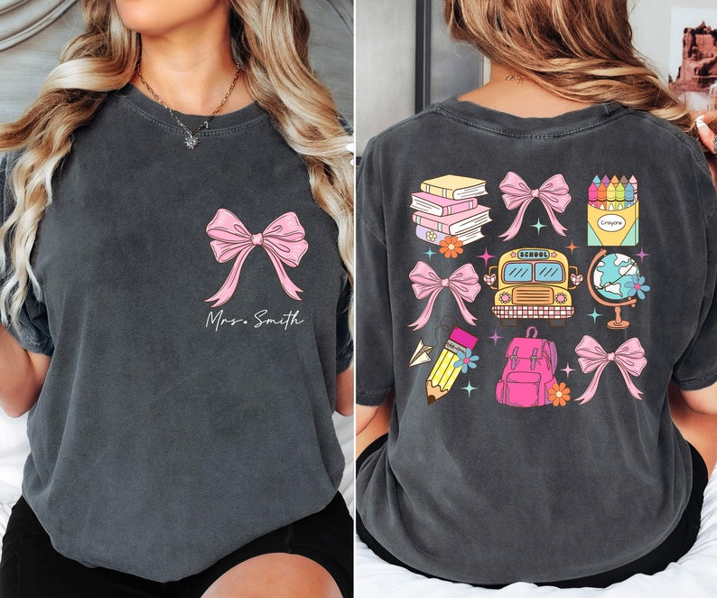 Op de afbeelding: Twee grijze T-shirts met een roze strik en de tekst "Mrs. Smith" op de voorkant en een schoolbus, rugzak, globe, boeken, potloden, kleurpotloden en strikken op de achterkant.
