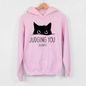 May include: A pink hoodie with a black cat peeking out from the top and the text "Judging You Silently" printed below.