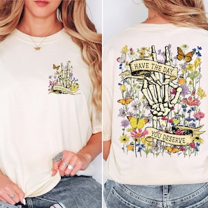 K&ouml;nnte beinhalten: Ein wei&szlig;es T-Shirt mit einem Blumendesign und einer Skeletthand, die ein Peace-Zeichen macht. Der Text "Have the day you deserve" ist auf einem Banner geschrieben.
