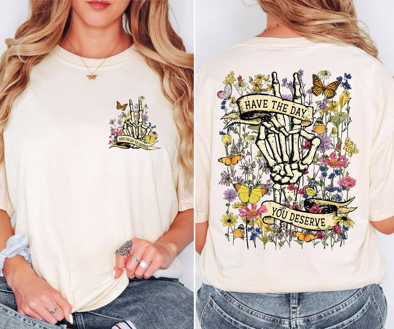 K&ouml;nnte beinhalten: Ein wei&szlig;es T-Shirt mit einem Blumendesign und einer Skeletthand, die ein Peace-Zeichen macht. Der Text "Have the day you deserve" ist auf einem Banner geschrieben.