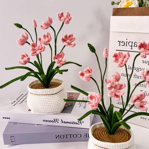 Pode incluir: Dois arranjos de flores de crochê em vasos brancos. As flores rosa têm caules e folhas verdes. Os vasos são brancos com uma textura tecida e estão em cima de livros. O fundo inclui um saco de papel com texto.