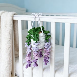 May include: A white crochet hanging planter with green leaves and purple flowers. The planter is suspended from a white crib rail. The flowers cascade downwards, adding a touch of color and texture to the nursery decor. The overall theme is handmade and whimsical.