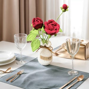 May include: A crocheted bouquet of red roses in a white crocheted vase with a brown tag that says "Hand Made". The vase is sitting on a grey placemat on a white table.