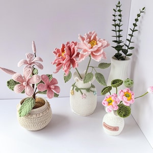 May include: A collection of handmade crochet flowers in various shades of pink and yellow, displayed in small, decorative vases. The flowers include peonies and other floral designs, with green leaves and stems. The vases are white and crocheted.