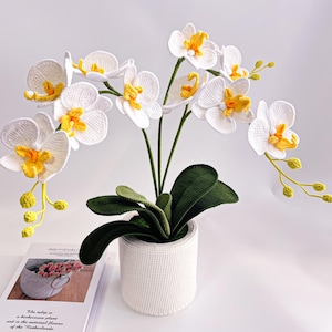 May include: A crocheted white orchid plant with yellow centers and green stems and leaves. The plant is in a white pot. The plant is on a white surface.