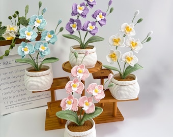 May include: Crocheted orchid plants in white pots, displayed on a wooden tiered stand. The orchids feature various colors, including blue, purple, pink, and white, with yellow centers and green leaves. The pots have a textured design.