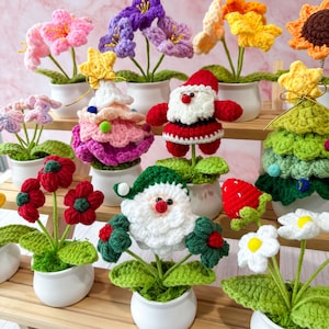 May include: A collection of colorful crochet flowers and holiday-themed decorations in white pots. The display includes pink, purple, yellow, and red flowers, along with a Christmas tree, Santa Claus, and a strawberry. The items are arranged on wooden shelves.