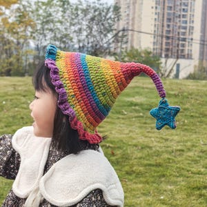 May include: A colorful, handmade crochet hat with a rainbow pattern. The hat has a ruffled brim and a long, curved tip with a blue star charm. The hat is worn by a person.