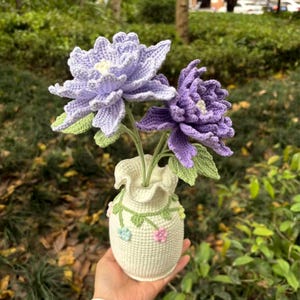 May include: Two crocheted purple flowers in a white crocheted vase with green vines and colorful flowers.