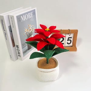 May include: A crocheted poinsettia plant with vibrant red petals and green leaves sits in a white pot. The plant is placed on a white surface next to two books and a wooden calendar displaying the number 25.