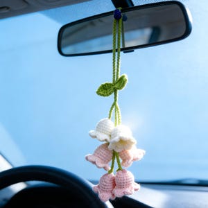 Puede incluir: Un adorno para coche hecho a mano con flores de ganchillo blancas y rosas, suspendido de un cordón verde. El adorno incluye un acento de hoja verde y cuelga de un espejo retrovisor contra un cielo azul brillante.