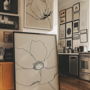 May include: A large framed art print featuring a black line drawing of a flower. The artwork is displayed on a wooden floor, with other framed art pieces visible on the wall. The color palette is neutral, with black and white dominating the image.