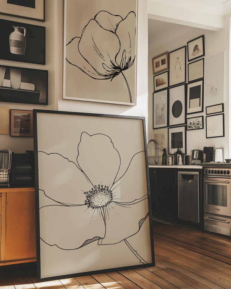 May include: A large framed art print featuring a black line drawing of a flower. The artwork is displayed on a wooden floor, with other framed art pieces visible on the wall. The color palette is neutral, with black and white dominating the image.