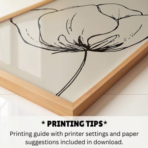 May include: A framed art print featuring a black and white floral illustration. The artwork is a minimalist line drawing of a flower against a cream-colored background, encased in a light brown wooden frame.