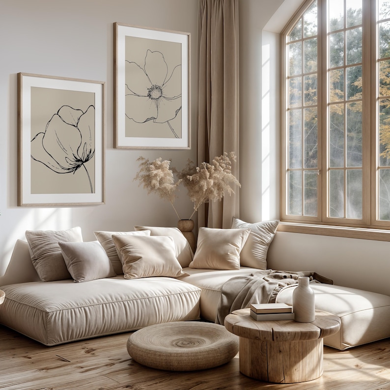 May include: Two framed botanical art prints featuring black line drawings of flowers on a beige background. The prints hang above a beige sectional sofa with several pillows. A wooden coffee table and a round ottoman are in front of the sofa.