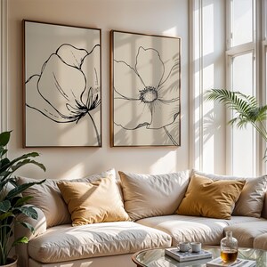 May include: Two framed floral art prints with black line drawings on a cream background. The artwork is displayed above a beige sectional sofa with mustard-colored throw pillows. A glass coffee table and potted plants complete the living room setting.