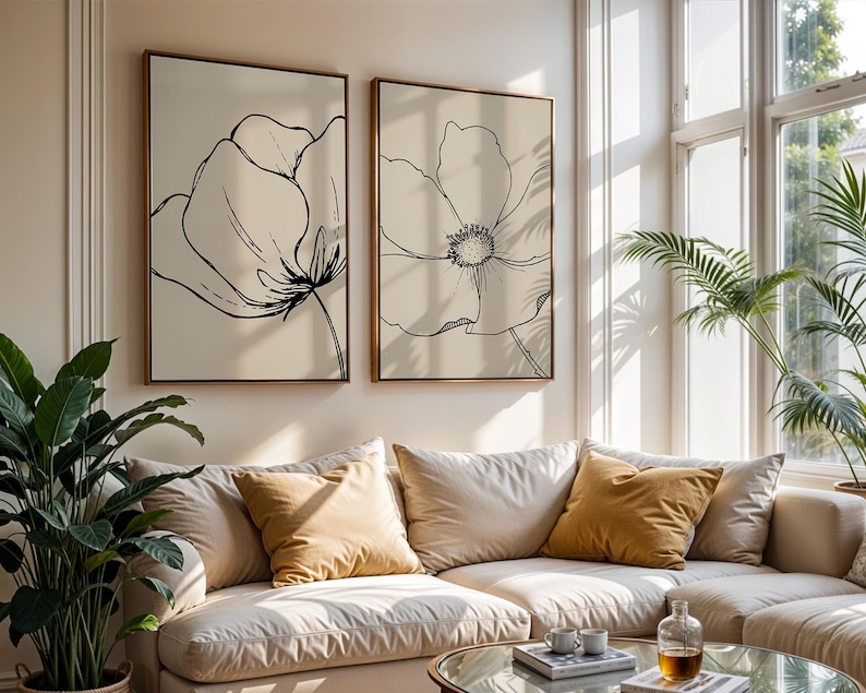 May include: Two framed floral art prints with black line drawings on a cream background. The artwork is displayed above a beige sectional sofa with mustard-colored throw pillows. A glass coffee table and potted plants complete the living room setting.
