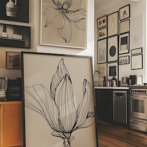 May include: A large framed artwork featuring a black and white line drawing of a magnolia flower. The artwork is leaning against a wall in a room with other framed art pieces and a kitchen area. The artwork is displayed on a light beige background.