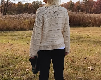 Könnte beinhalten: Ein cremefarbener Häkelpullover mit einem strukturierten Stichmuster. Der Pullover hat lange Ärmel und eine lockere Passform.
