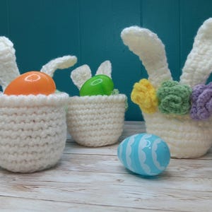 May include: Three white crocheted bunny baskets with floppy ears. The baskets are filled with plastic Easter eggs in orange, green, and blue. The baskets are decorated with crocheted flowers in yellow, green, and purple.