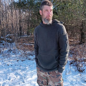 Peut inclure: Un sweat à capuche gris foncé avec une poche kangourou et des coudières renforcées. La personne porte un pantalon de camouflage et se tient dans une zone boisée et enneigée. Le sweat semble être fait d'une matière tricotée épaisse.