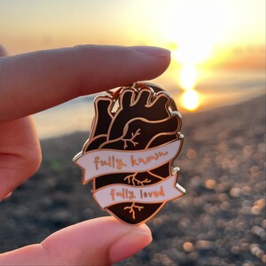 May include: Gold-tone heart-shaped enamel pin with a black anatomical heart design. The pin has two white banners with the text "fully known" and "fully loved" in black.