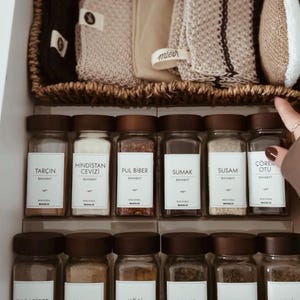 May include: A kitchen drawer organized with spice jars and woven storage baskets. The spice jars have dark brown lids and white labels with Turkish names. The woven basket contains beige and black textiles.