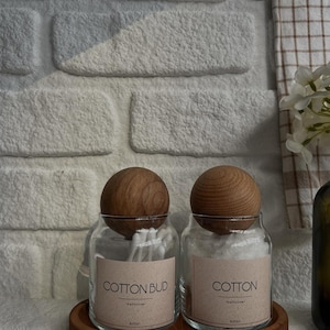 May include: Two clear glass jars with wooden lids, labeled "COTTON BUD" and "COTTON", sit on a wooden tray. The jars are filled with cotton swabs and cotton balls. The background features a white brick wall and a checkered towel.