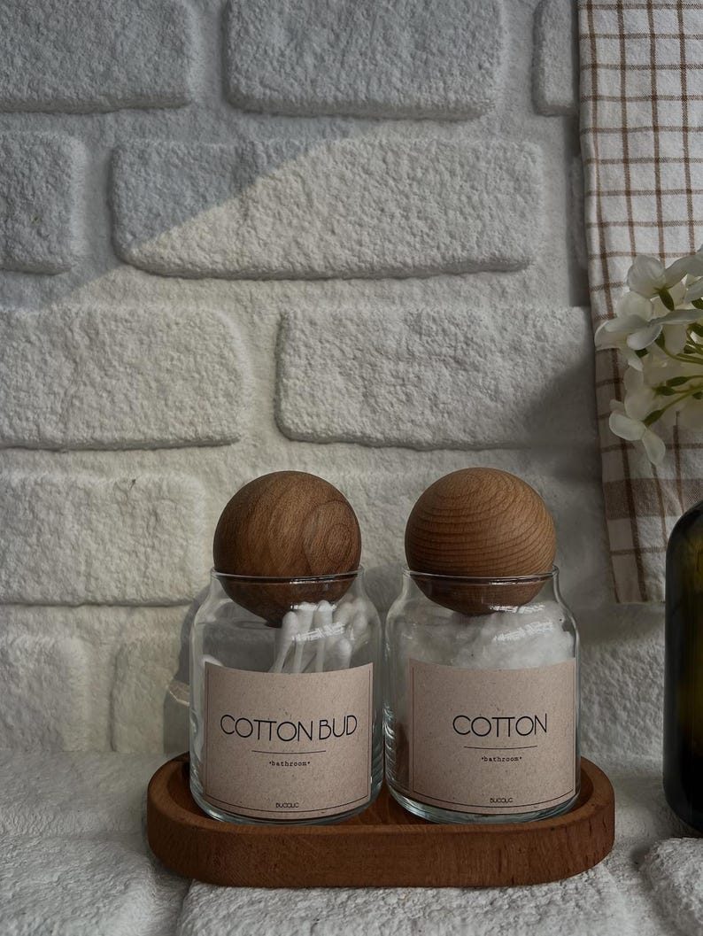 May include: Two clear glass jars with wooden lids, labeled "COTTON BUD" and "COTTON", sit on a wooden tray. The jars are filled with cotton swabs and cotton balls. The background features a white brick wall and a checkered towel.