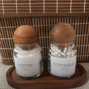 May include: Two glass jars with wooden lids and labels. The jar on the left is labelled "COTTON" and the jar on the right is labelled "COTTON BUD". Both jars are sitting on a wooden tray.