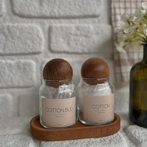 May include: Two clear glass jars with wooden lids, labeled "COTTON BUD" and "COTTON", sit on a wooden tray. The jars are filled with cotton swabs and cotton balls. The background includes a white brick wall and a vase with flowers.