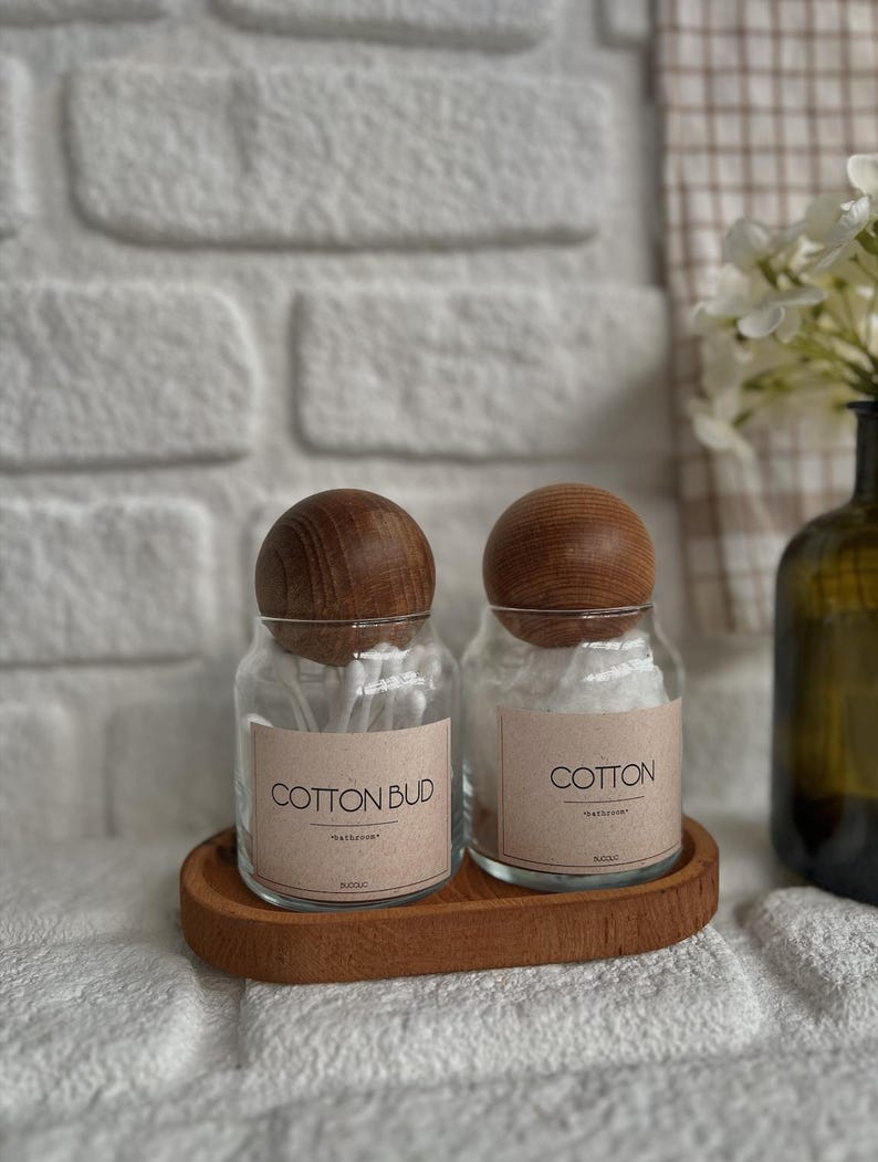 May include: Two clear glass jars with wooden lids, labeled "COTTON BUD" and "COTTON", sit on a wooden tray. The jars are filled with cotton swabs and cotton balls. The background includes a white brick wall and a vase with flowers.