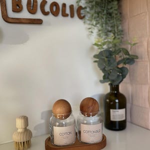 May include: Two clear glass jars with wooden lids labeled "COTTON" and "COTTON BUD" sit on a wooden tray. A wooden brush and a decorative wooden sign that says "BUCOLIC" are also visible. A green glass bottle with a plant is in the background.