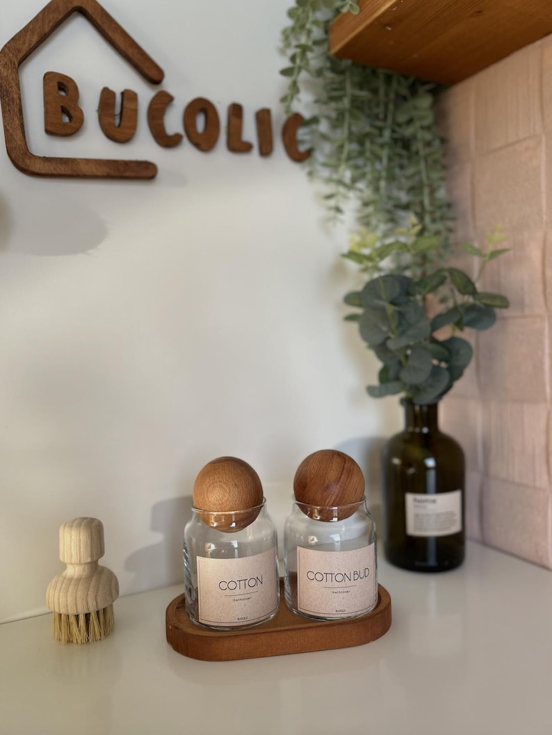 May include: Two clear glass jars with wooden lids labeled "COTTON" and "COTTON BUD" sit on a wooden tray. A wooden brush and a decorative wooden sign that says "BUCOLIC" are also visible. A green glass bottle with a plant is in the background.